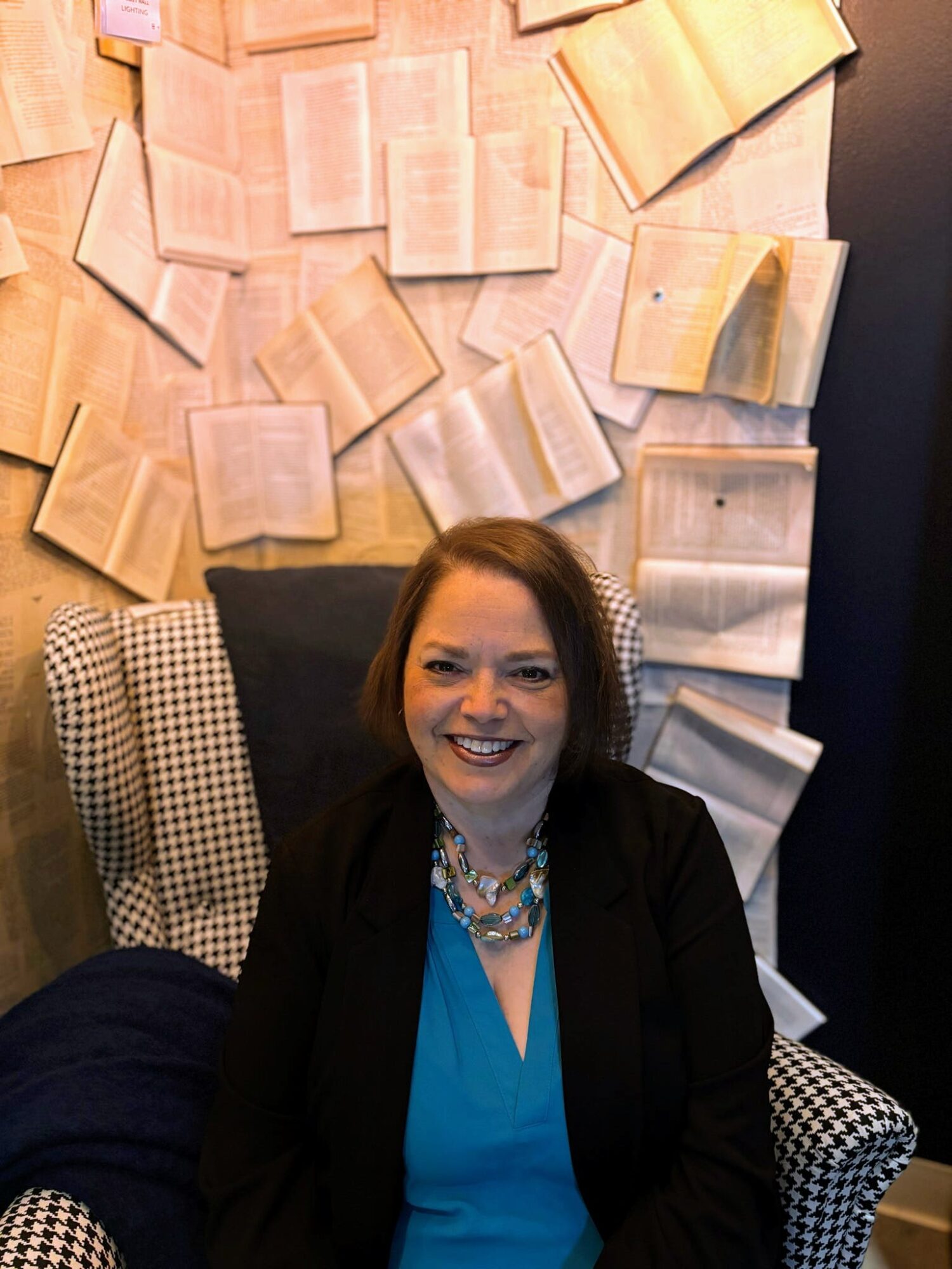 Woman with dark hair smiling, sitting in a patterned armchair, with open books on the wall behind her.