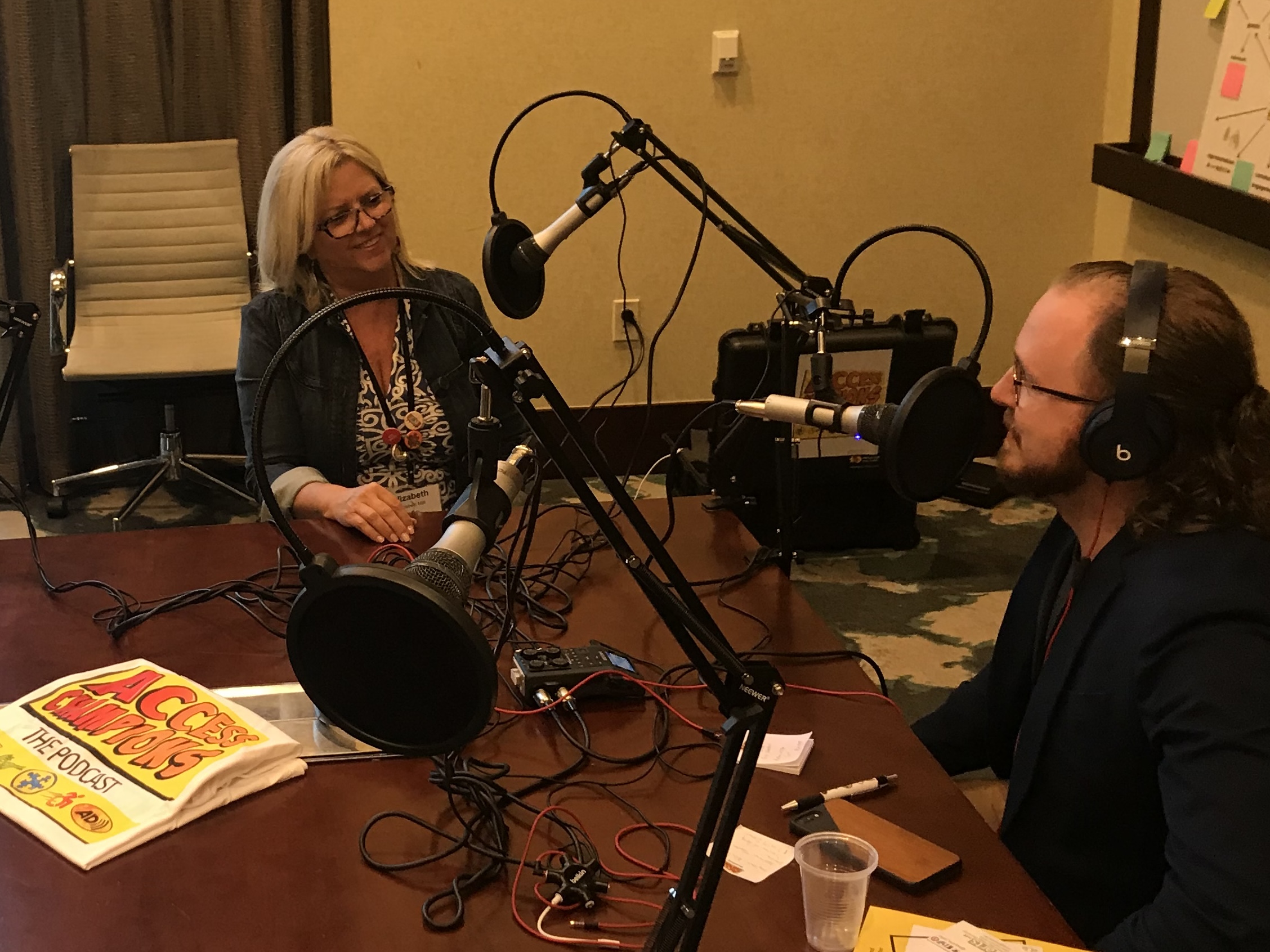 Two people sit at a table with microphones, headphones, and recording equipment in a room with yellow walls.