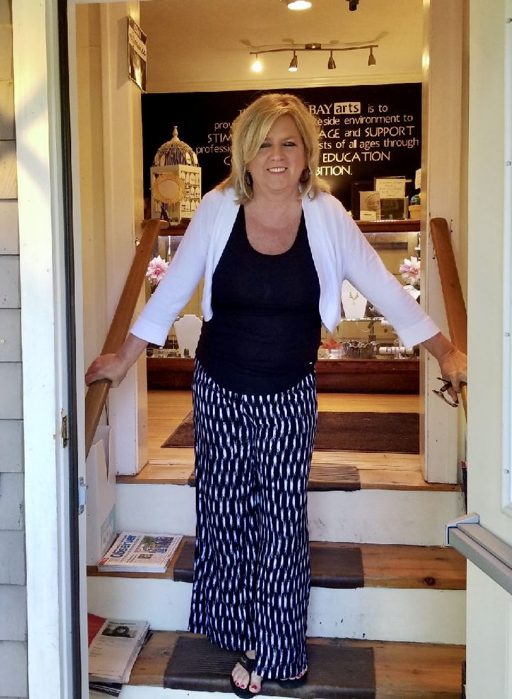 Woman with blonde hair smiling, standing in doorway, wearing a white cardigan, black top, and patterned pants, inside a store.