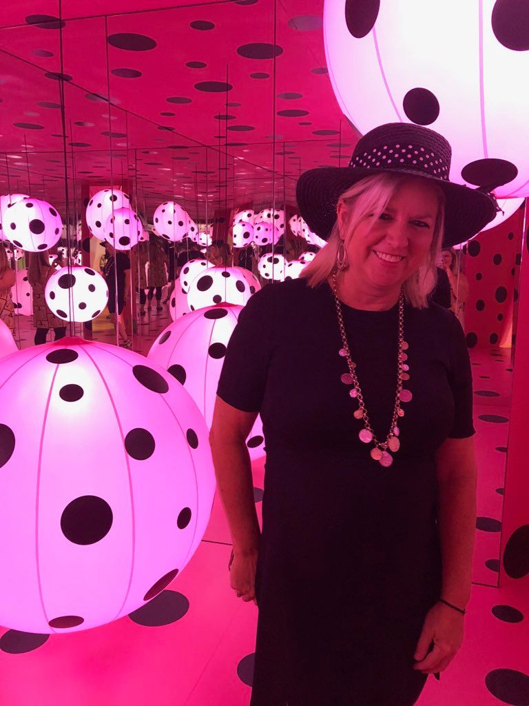Woman dressed in black with a hat and necklace, standing in front of a pink background with black polka dots.