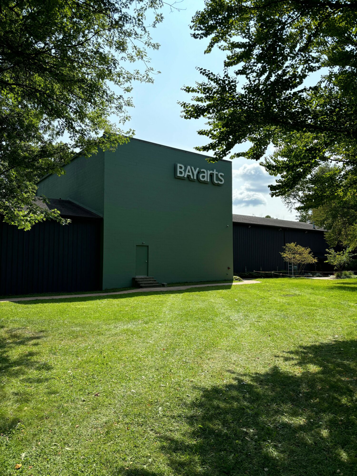 Green building with BayArts sign, surrounded by trees and grass, under a partly cloudy sky.