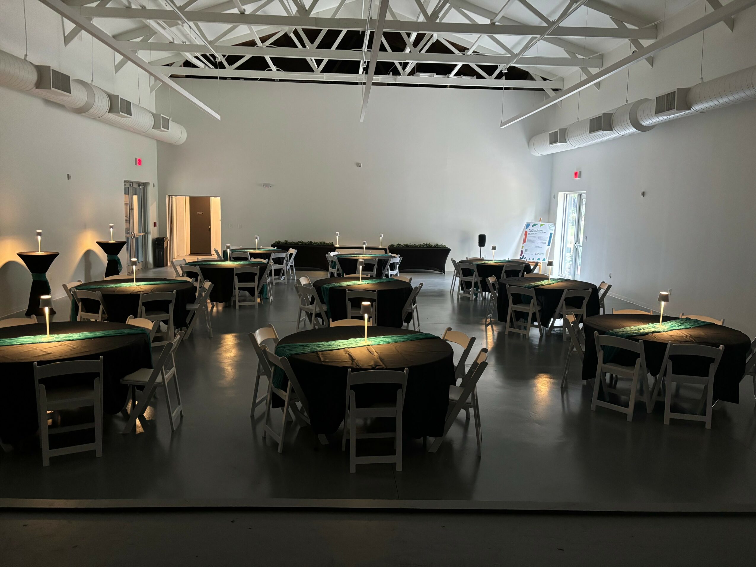 Room with round tables and chairs, lit by small lamps, with a high ceiling and white walls.