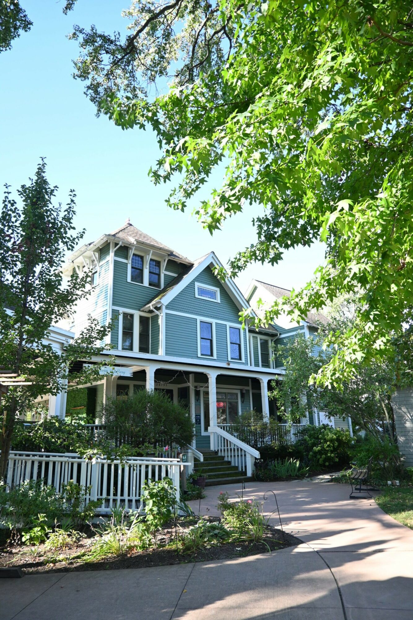 Three-story blue house with white trim, surrounded by trees and greenery, with a front porch and stairs leading to the sidewalk.