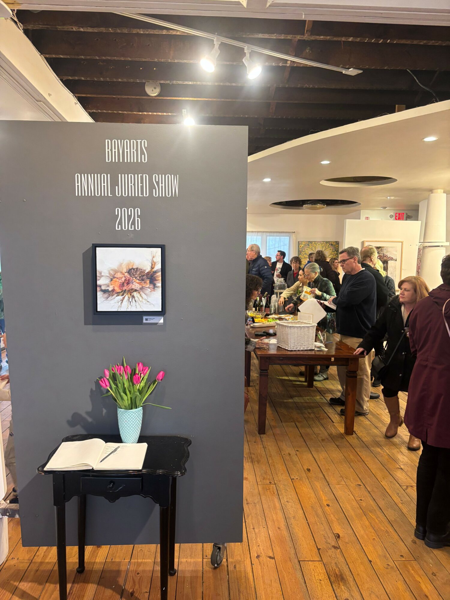 People gather in a room with wooden floors, a gray wall with framed art, and a table with a vase of pink tulips.