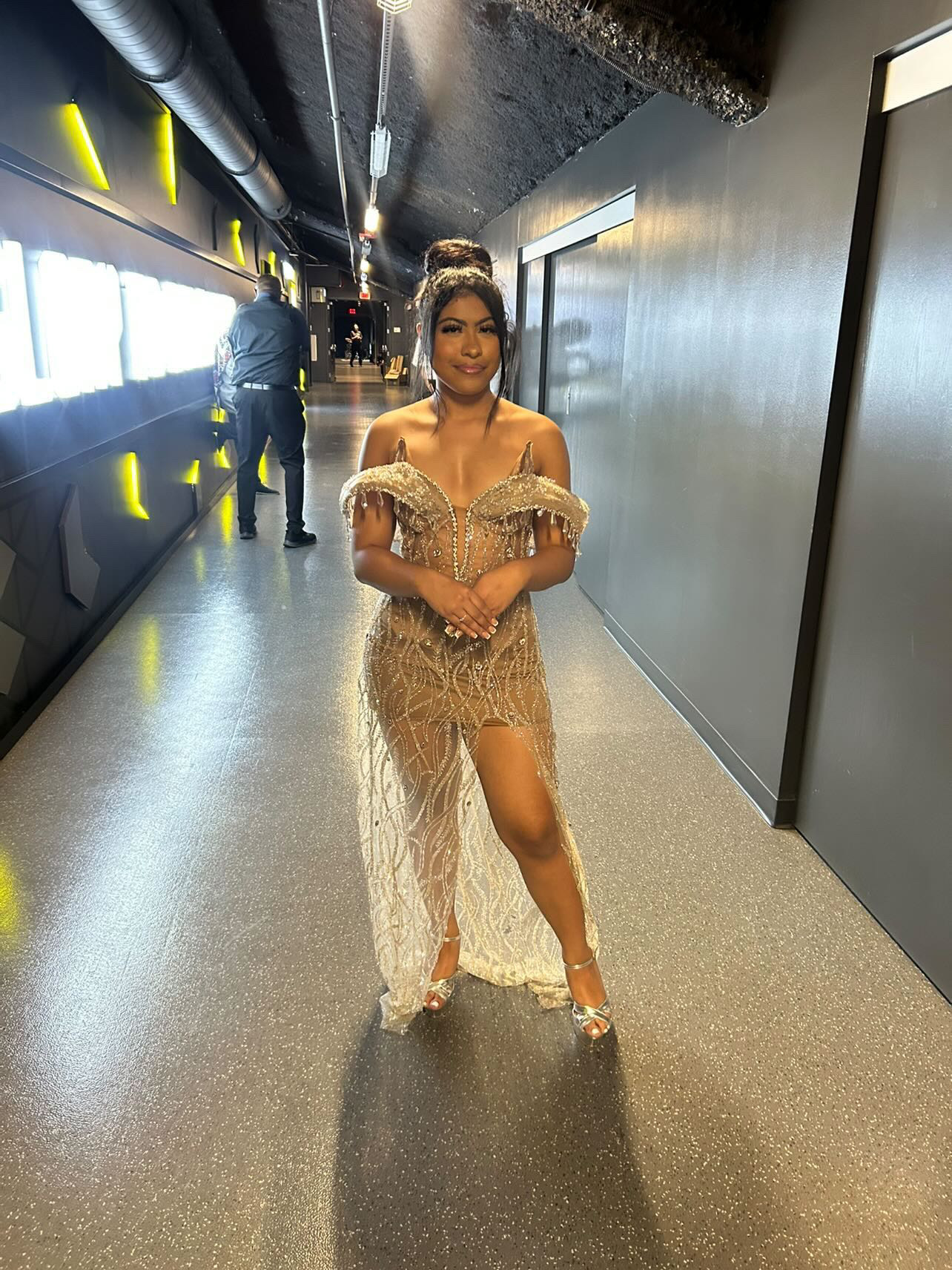 Woman in a sparkling dress standing in a modern hallway with yellow lights and large windows, smiling at the camera.