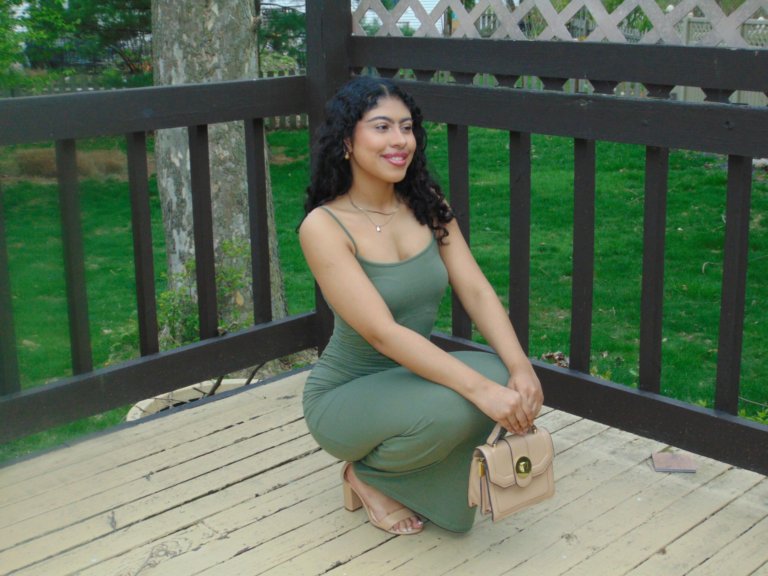 Young woman with curly hair kneeling on a wooden deck, smiling, holding a beige purse, wearing a green dress, outdoors.