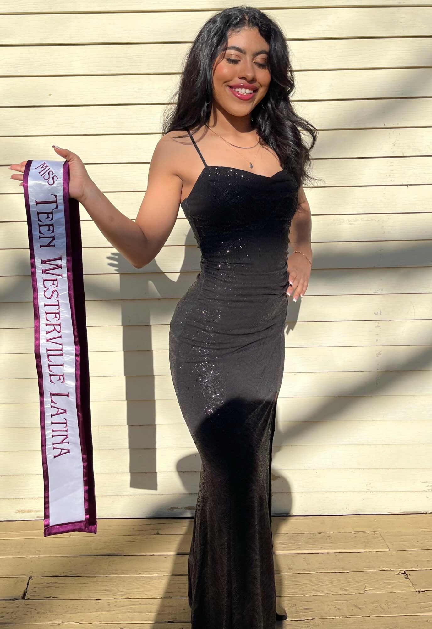 Young woman in a black dress holding a sash, standing outdoors against a beige wall, smiling.
