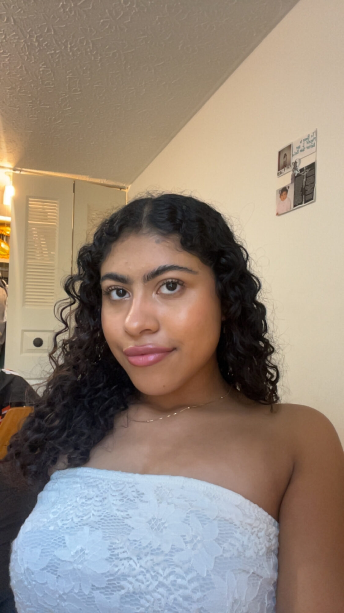 Young woman with curly hair wearing a strapless top, looking at the camera indoors.