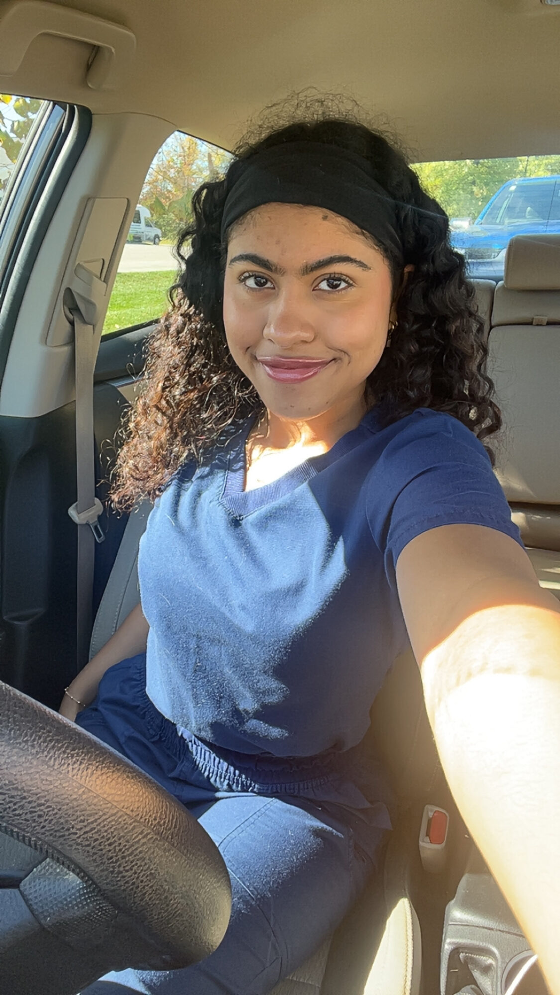 Young woman with curly hair and headband taking a selfie inside a car, smiling, sunlight outside.