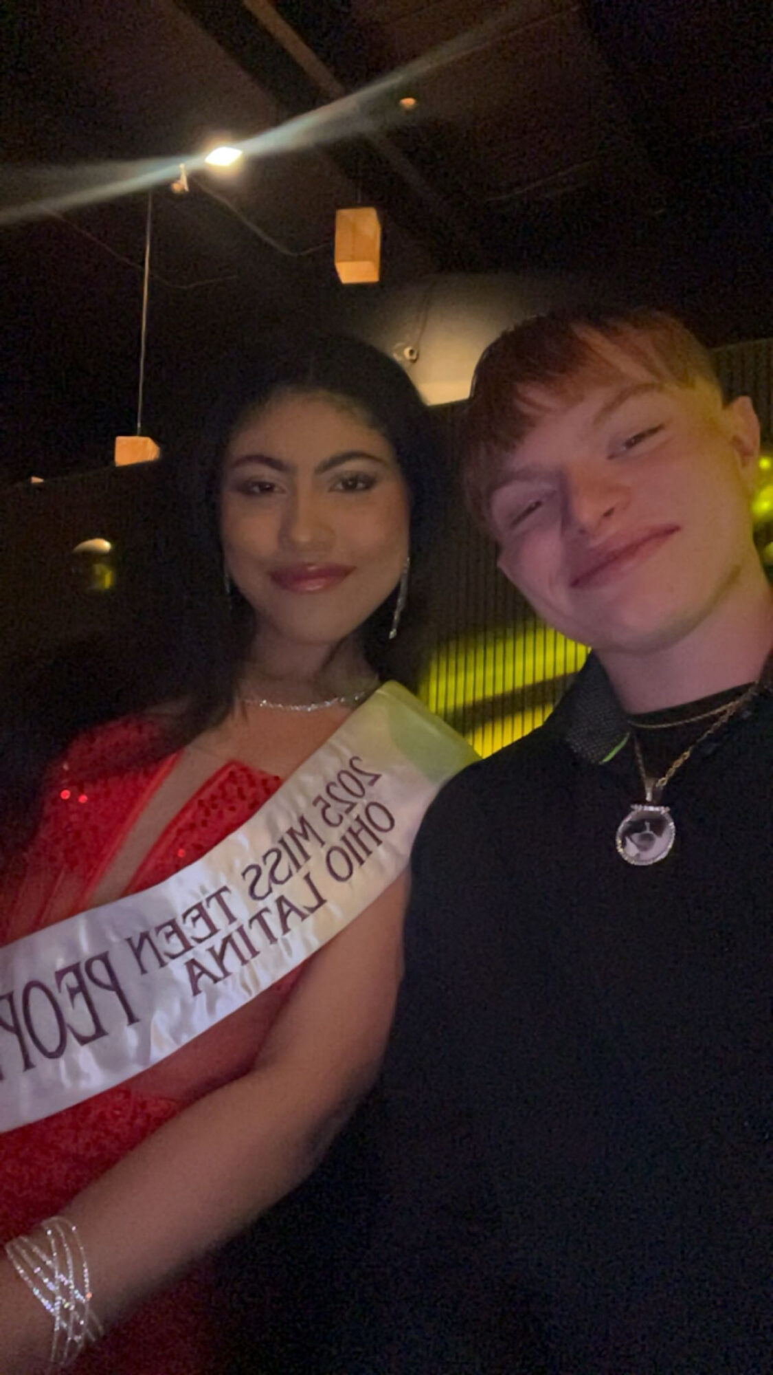 A young woman and a young man pose together in a dimly lit setting, smiling at the camera.