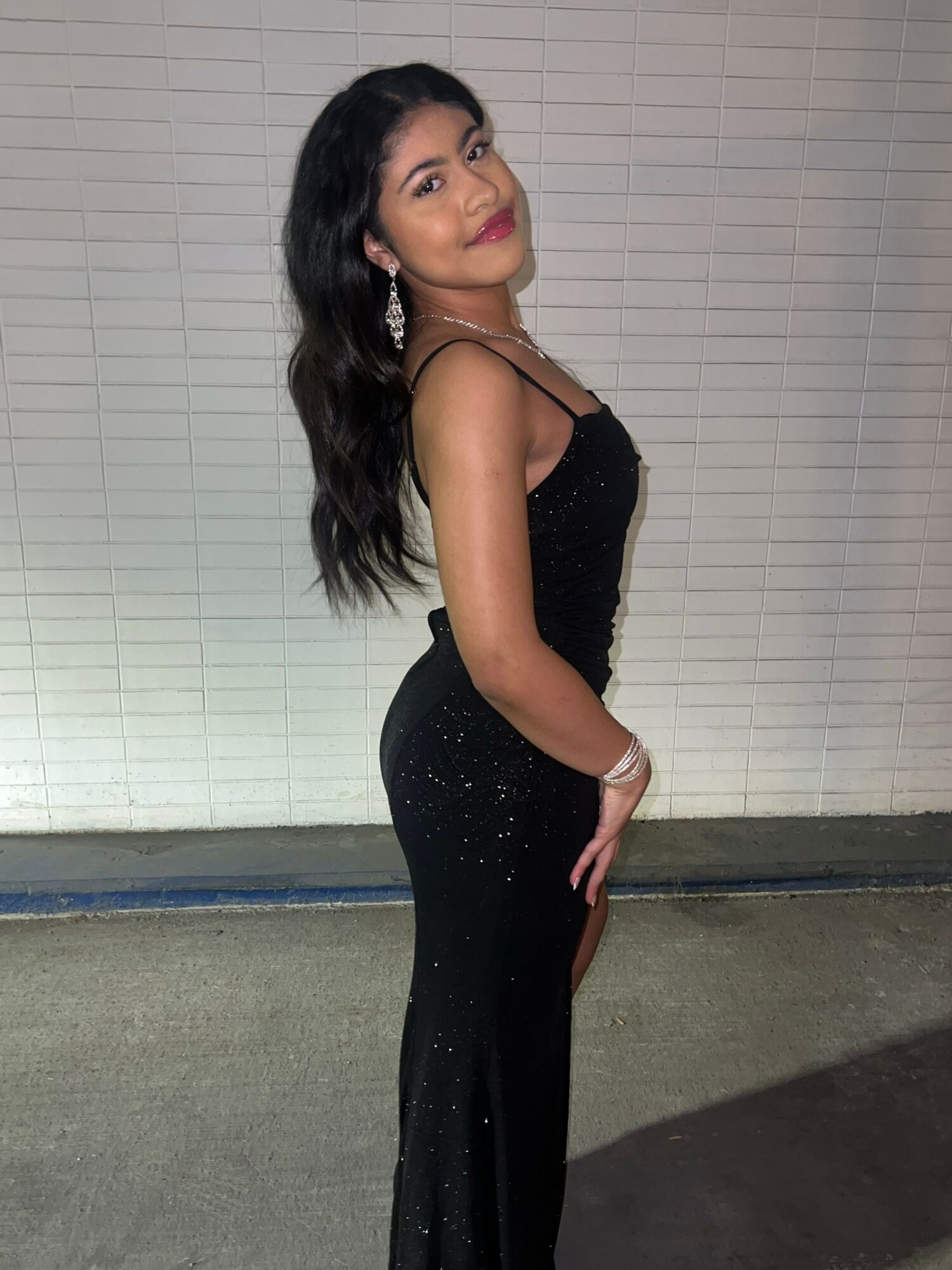 Woman in black dress standing sideways against a white tiled wall, looking at the camera with her hand on her hip.