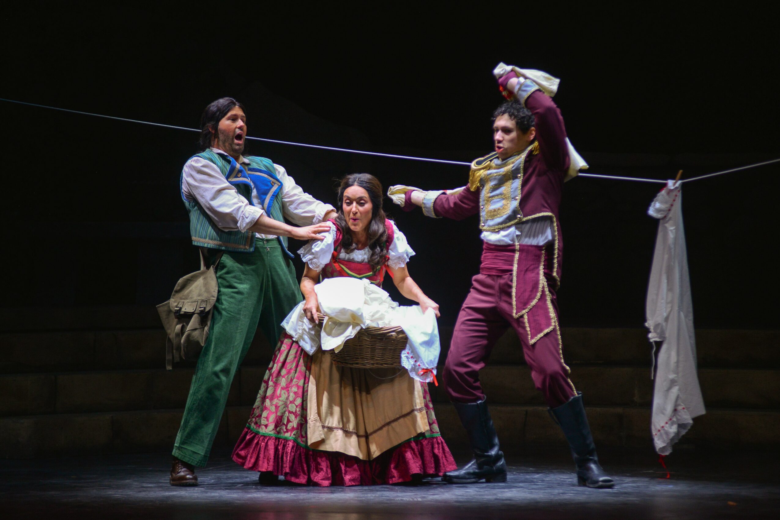 Three performers in colorful costumes on stage, with a clothesline and laundry in the background.