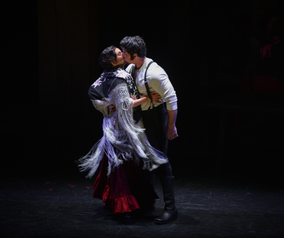Two people kiss on a dark stage, one wearing a fringed dress and the other in a white shirt with suspenders.