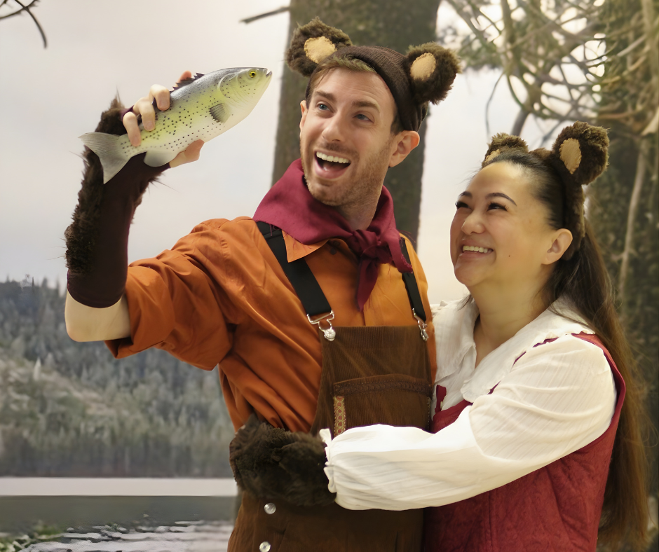 Two people wearing animal ears costumes, smiling, holding a fish outdoors near water and trees.