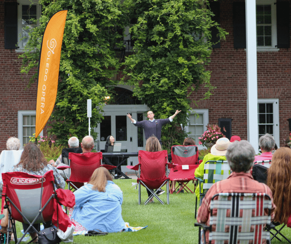 Person speaking outdoors to an audience seated on grass, with trees and a brick building in background.