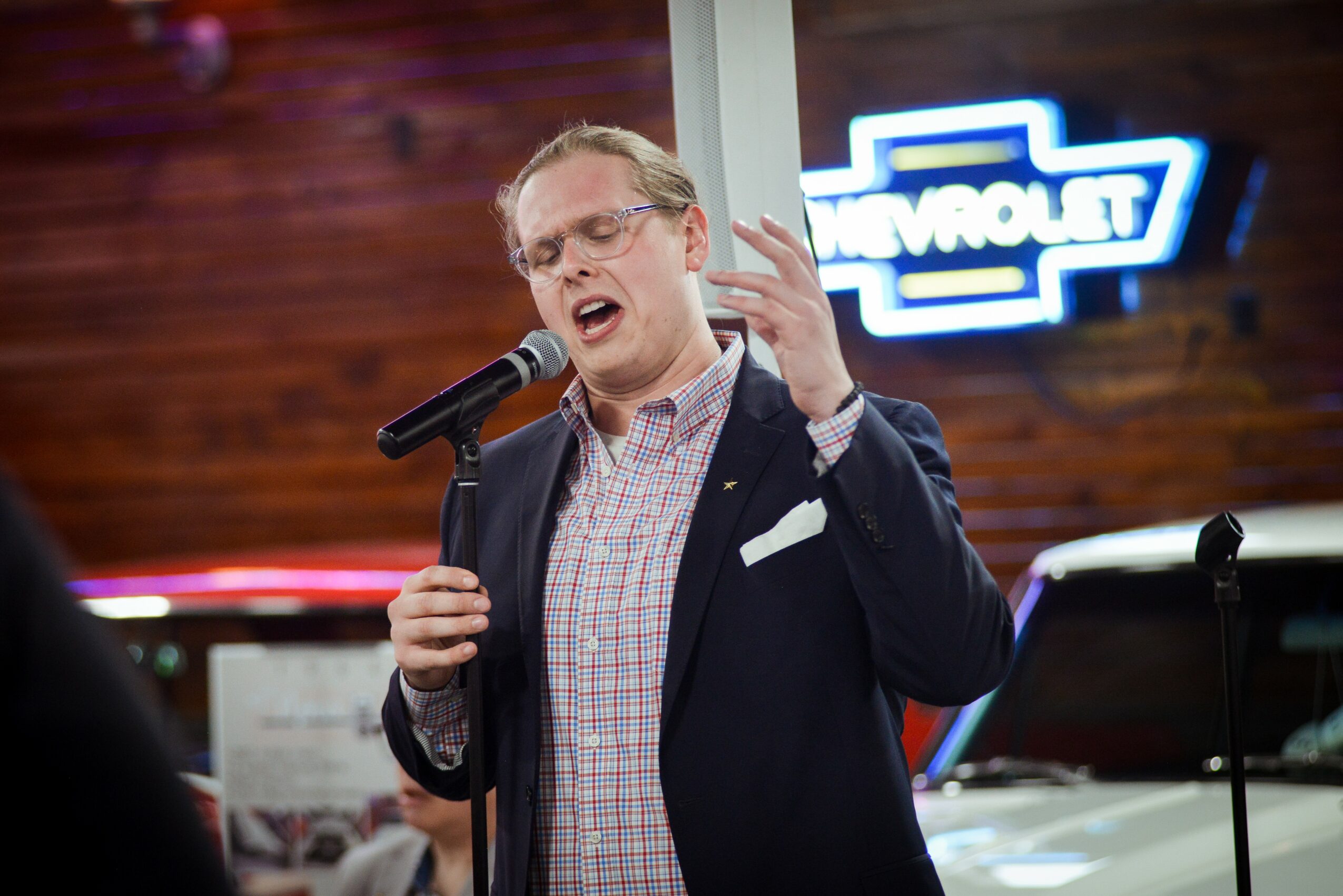 Person singing into a microphone with a neon sign in the background, gesturing with one hand, indoors.