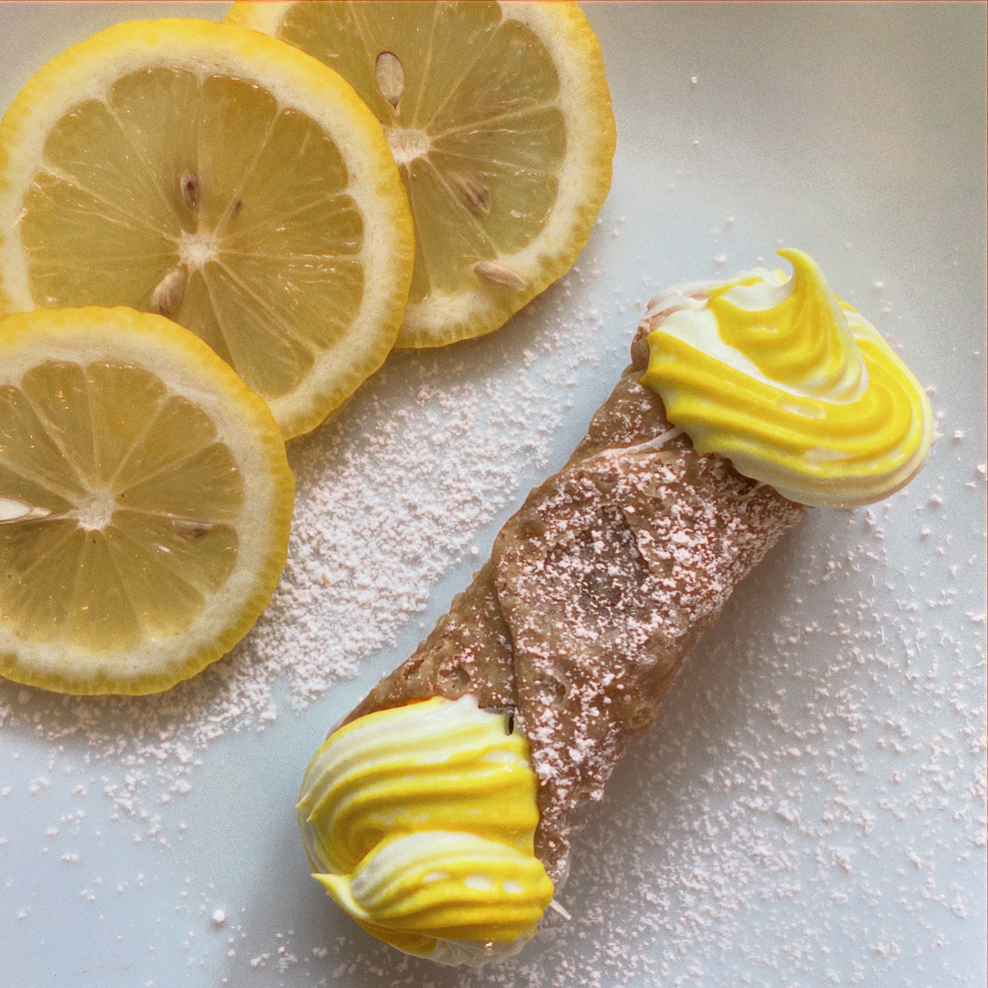 Lemon slices and a rolled pastry with yellow and white frosting on a white surface, dusted with powdered sugar.