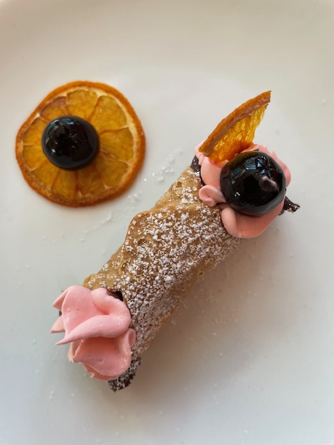A dessert resembling a fish with pink frosting, a slice of orange, and a cherry on top, dusted with powdered sugar.