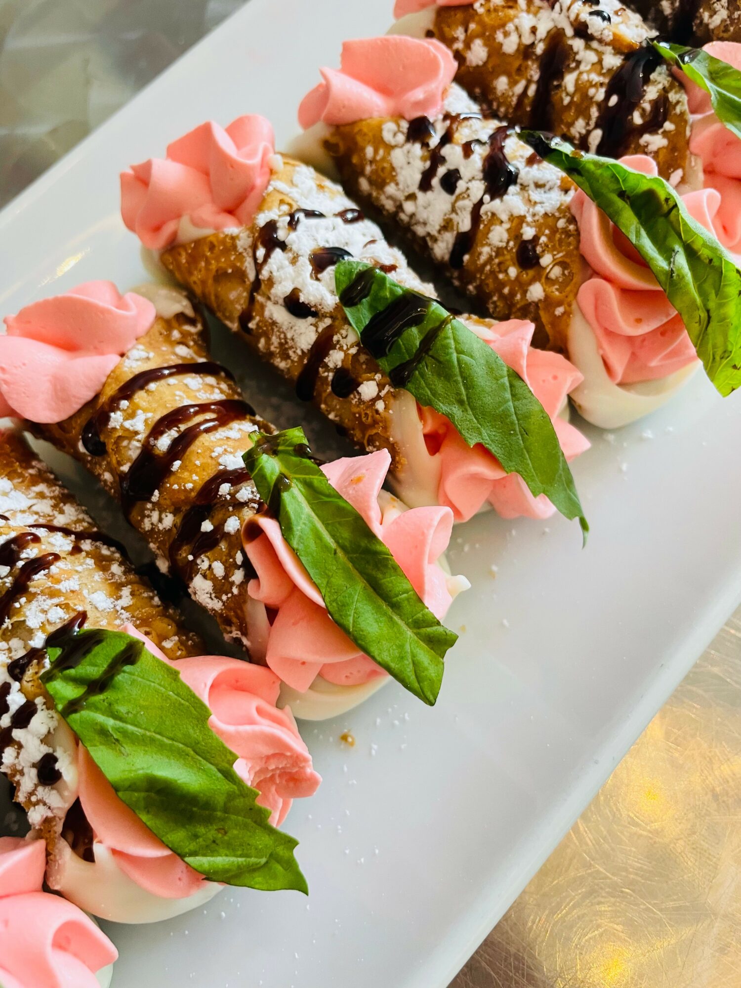 Plate with cannoli topped with pink frosting, green leaves, and drizzled chocolate, garnished with powdered sugar.