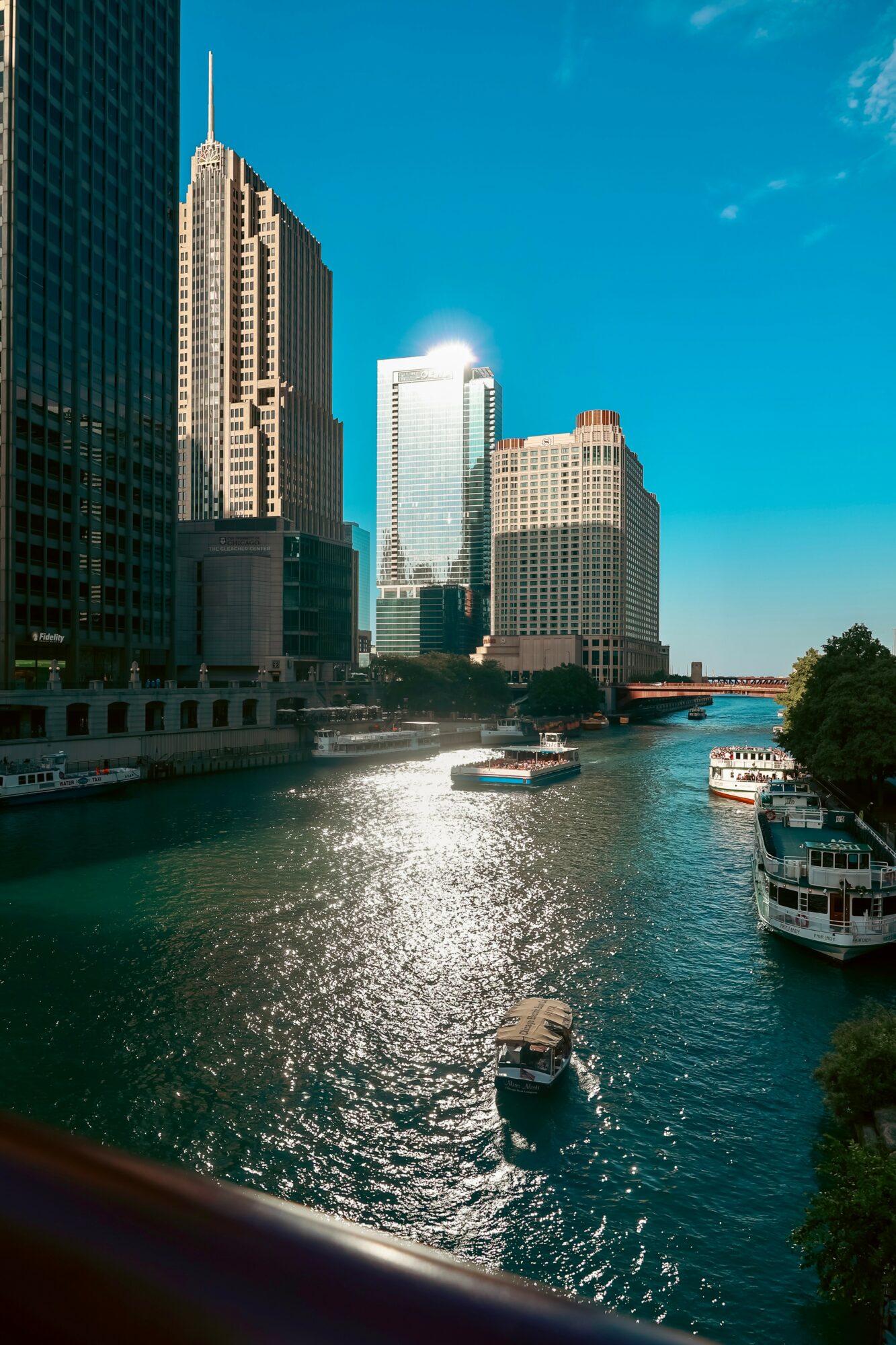 City skyline with tall buildings and boats on a river, under a blue sky with some clouds.