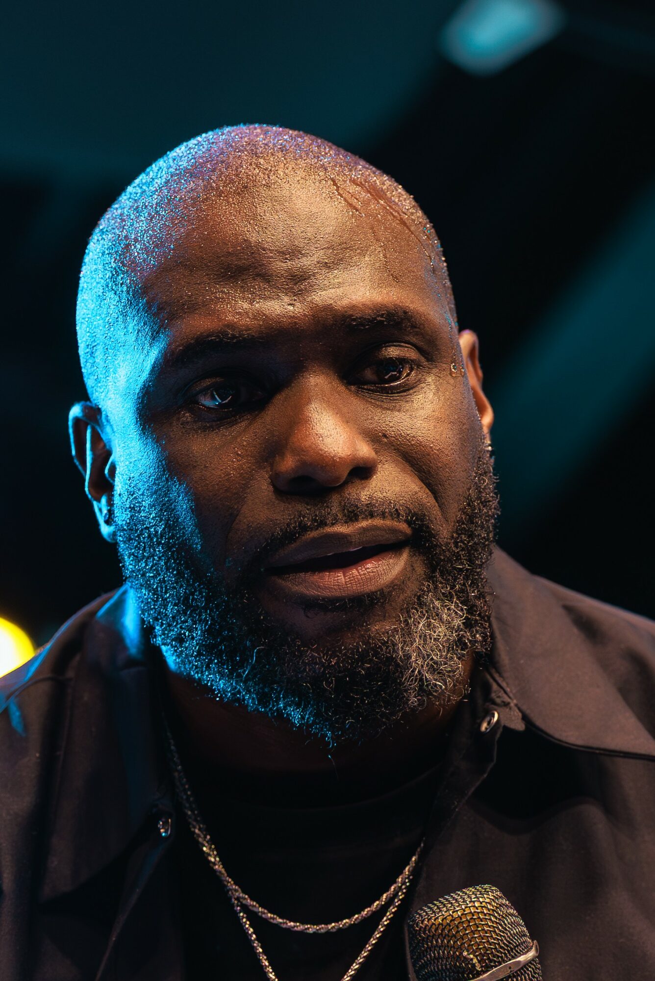 Close-up of a man with a beard and short hair, speaking into a microphone, with a dark background.