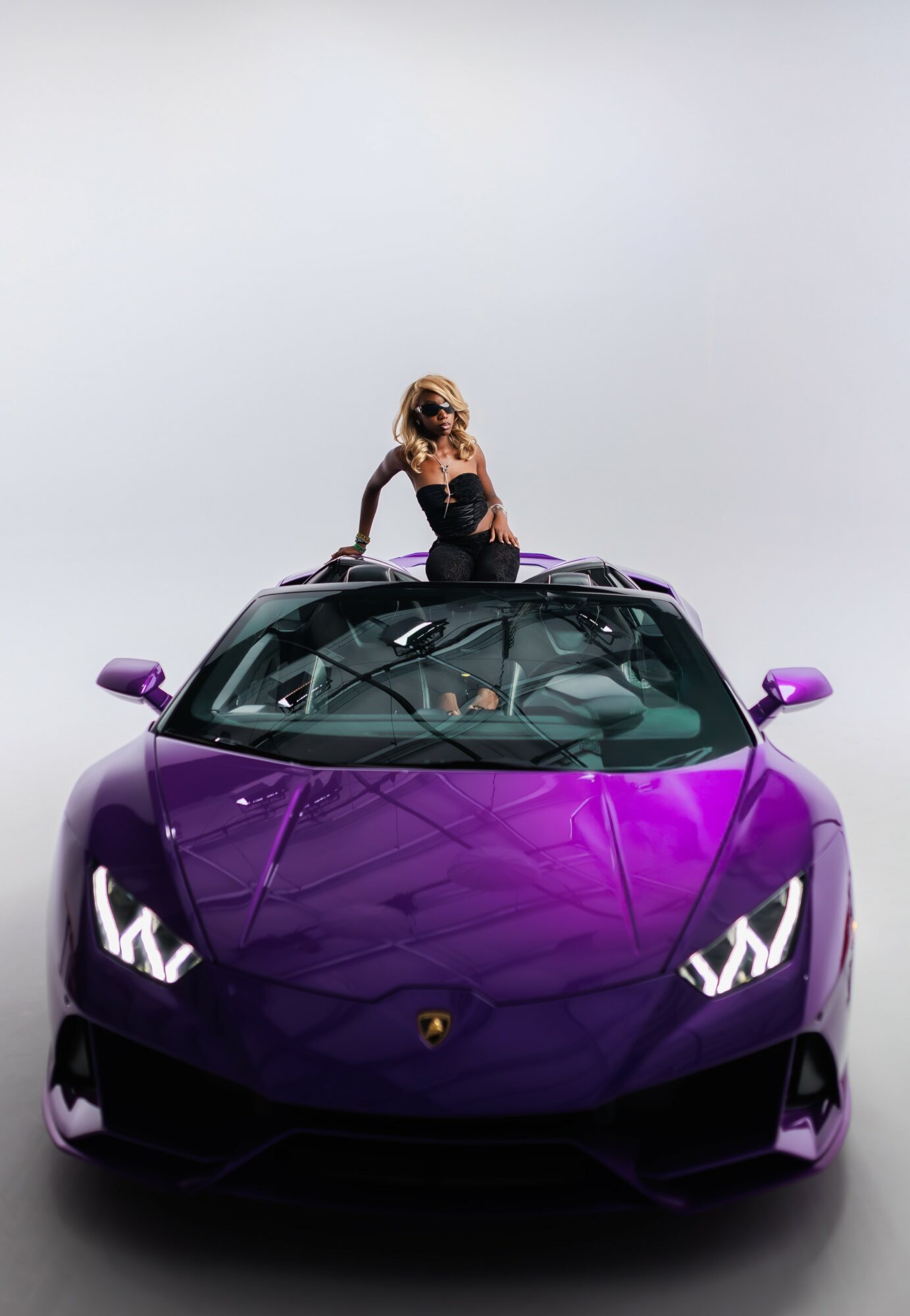 Woman with blonde hair sitting on a purple sports car, looking down, against a plain light background.