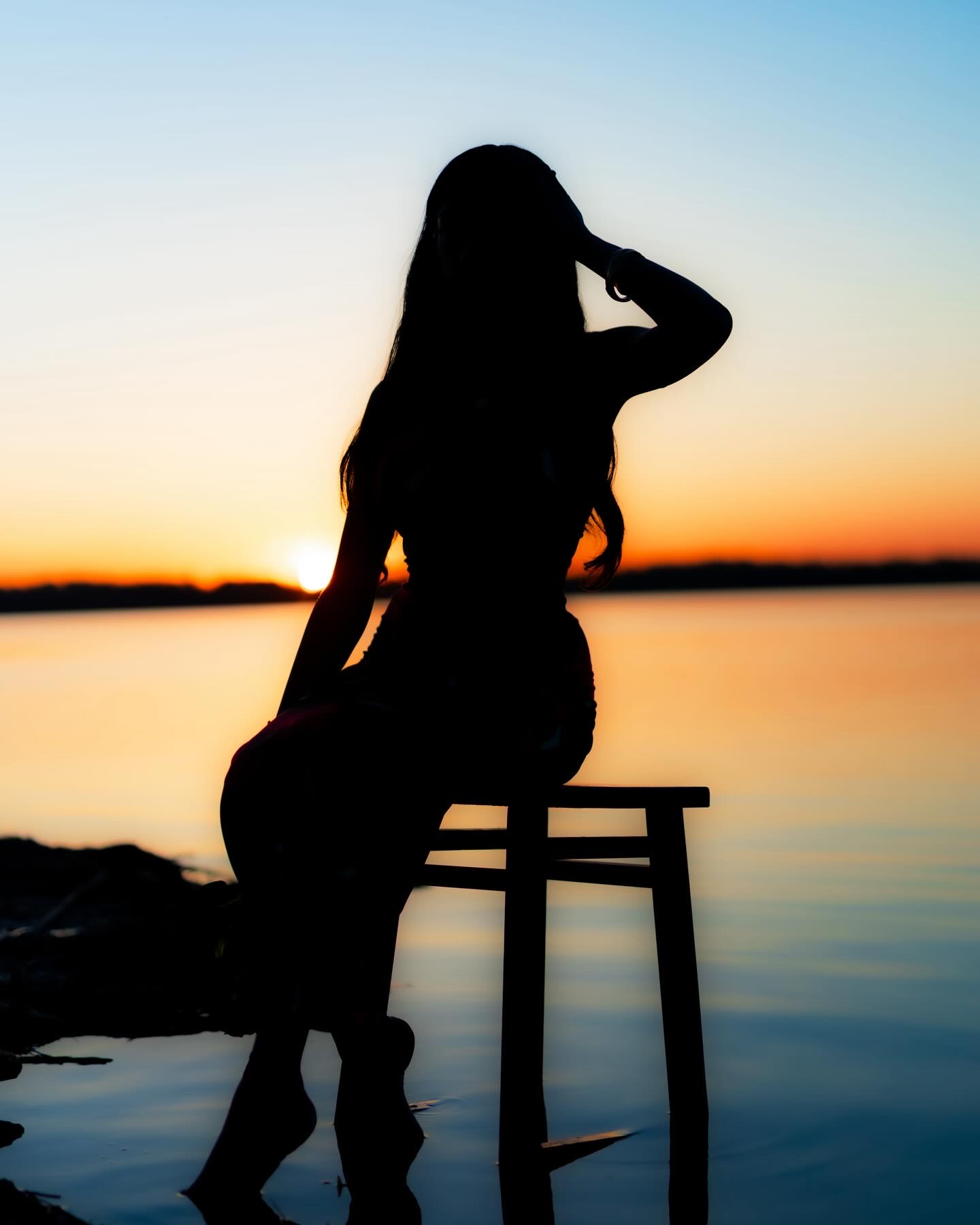 Silhouette of a woman sitting on a stool by water during sunset, with her hand near her face.
