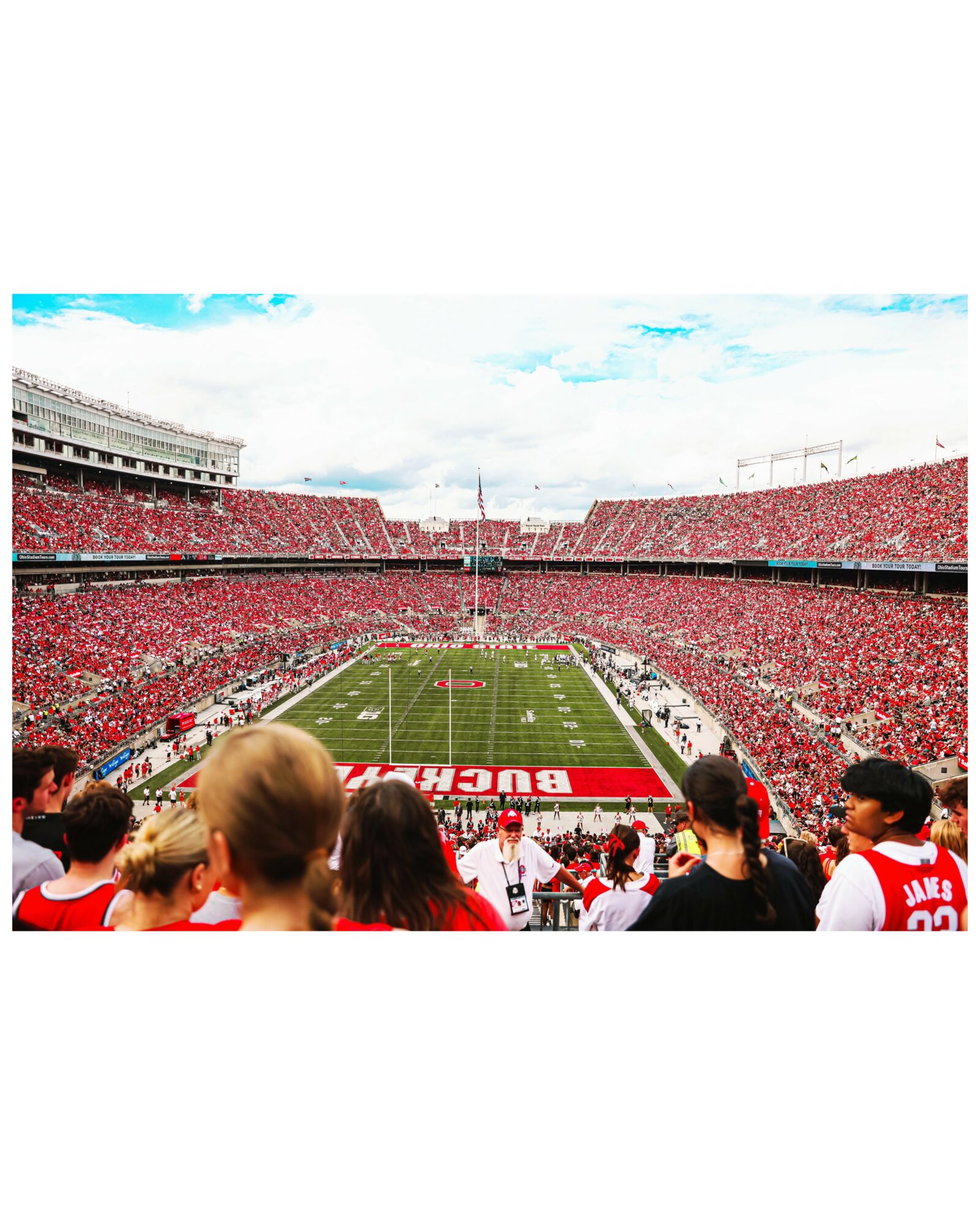 Football stadium filled with fans, view from the stands, field with team logo, sky with clouds, spectators in foreground.