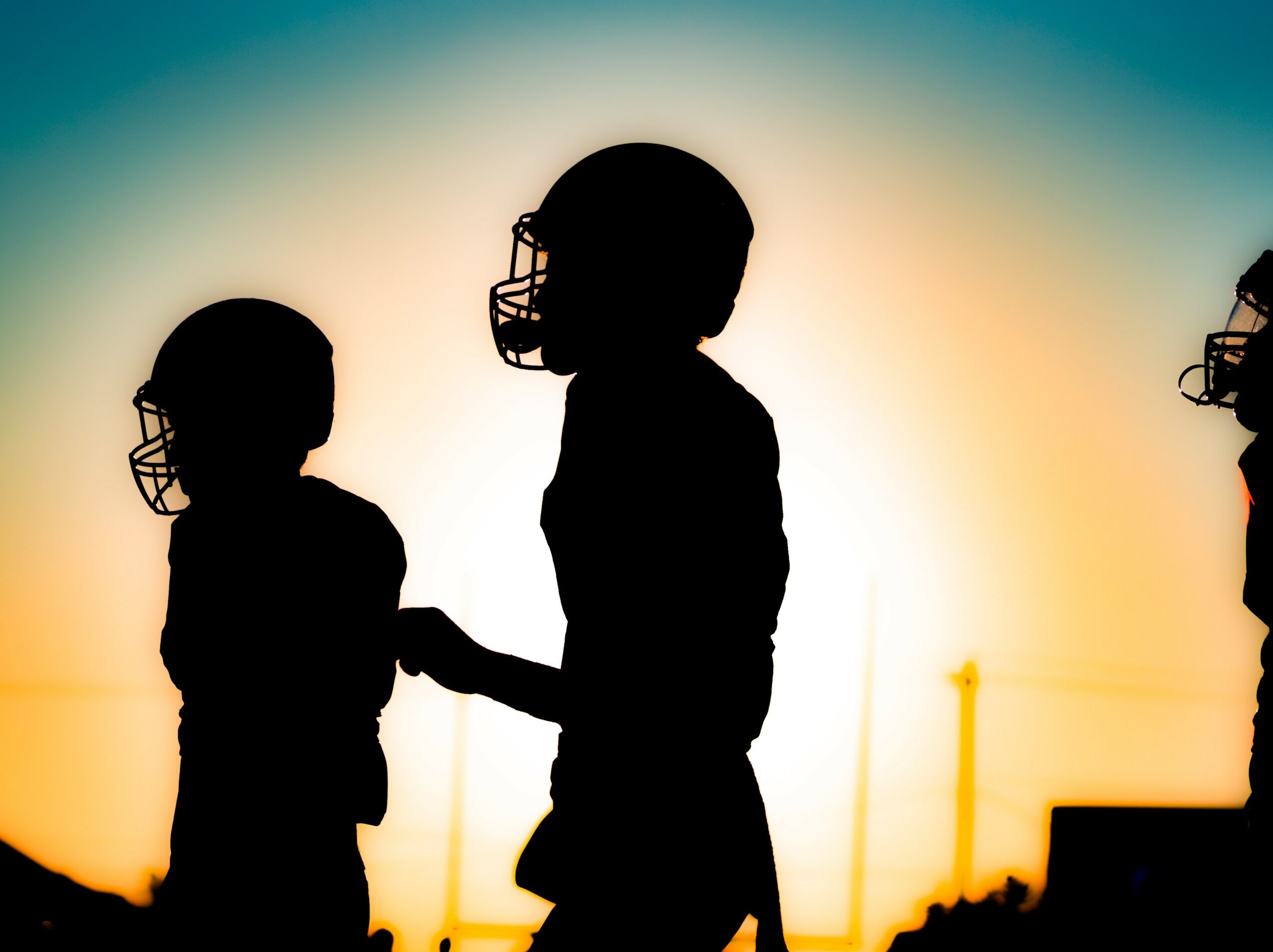 Silhouettes of two children wearing helmets, holding hands against a sunset sky with a gradient of blue and orange.