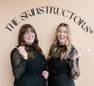 Two women smiling and holding party blowers, standing under a sign that reads 'THE SKINSTRUCTORS'.