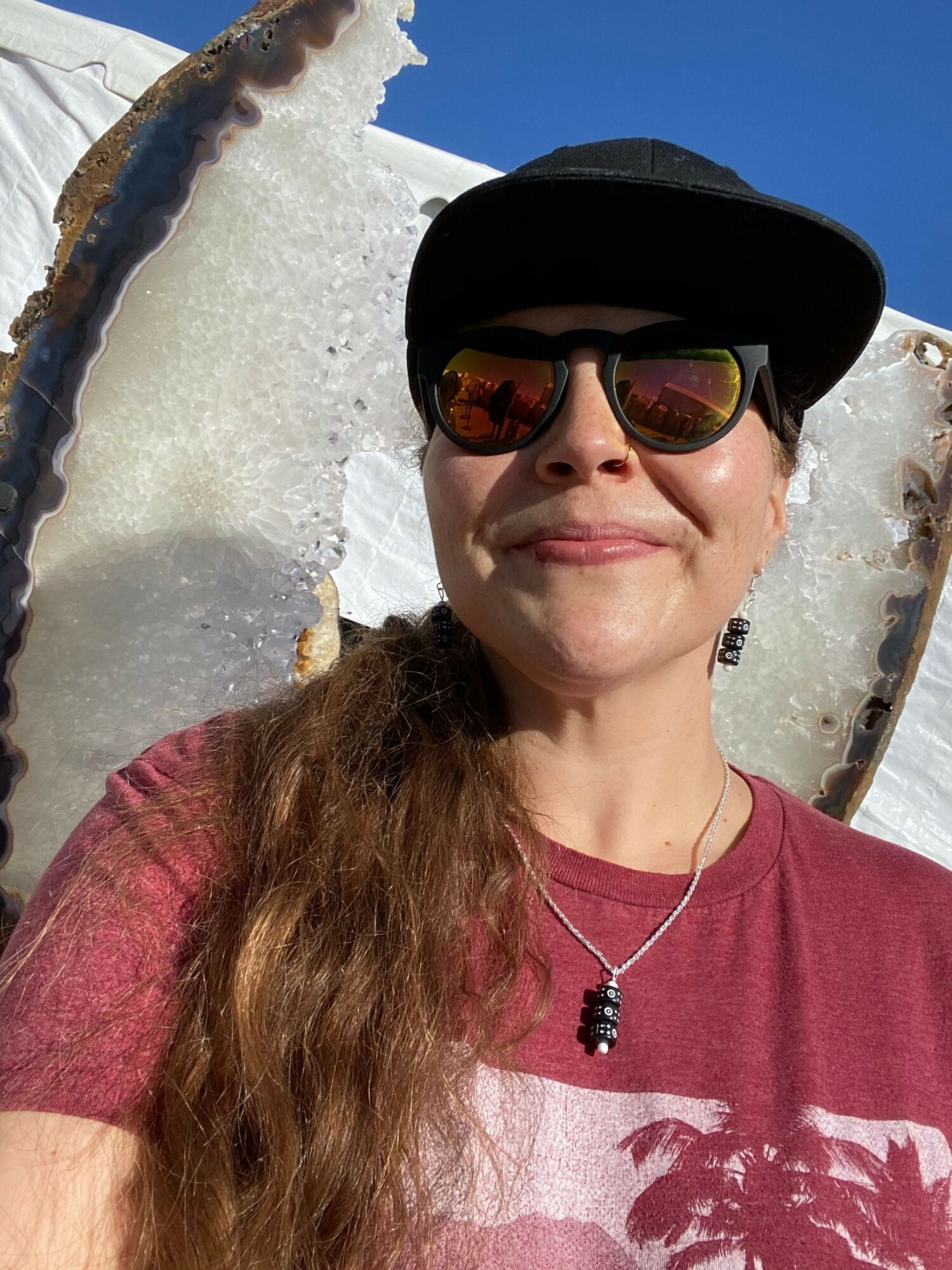 Person wearing sunglasses, a hat, and a necklace, smiling outdoors with a large shell and blue sky.