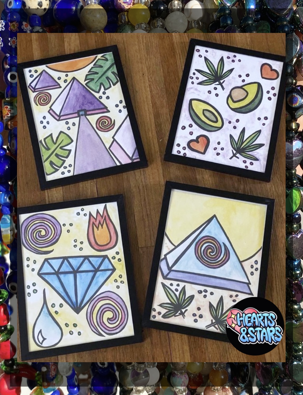 Four framed abstract illustrations with geometric shapes and patterns, placed on a wooden surface with colorful beads around.