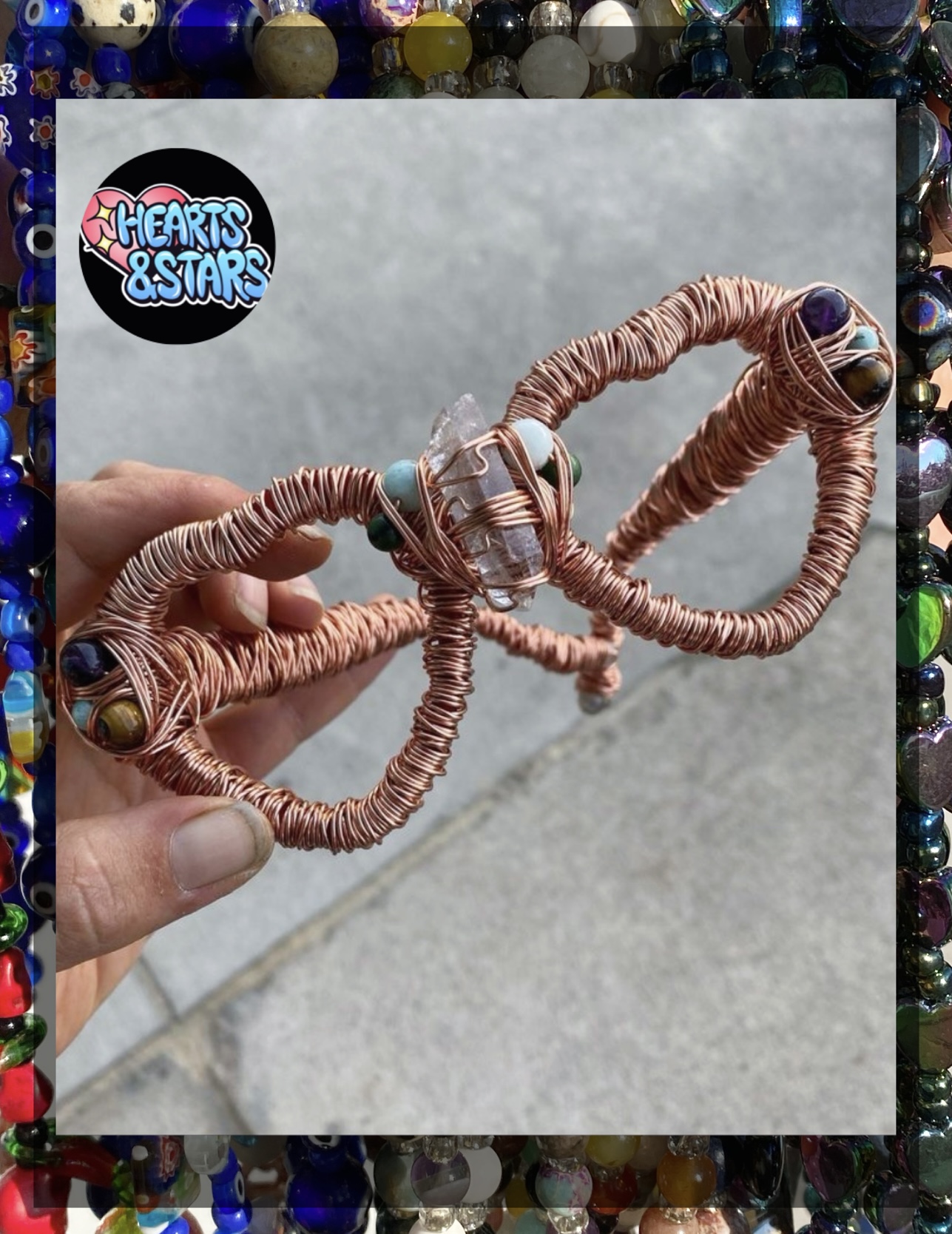 Hand holding a wire heart-shaped sculpture with beads, against a concrete background, surrounded by colorful beads. Word 'Hearts & Stars' logo.
