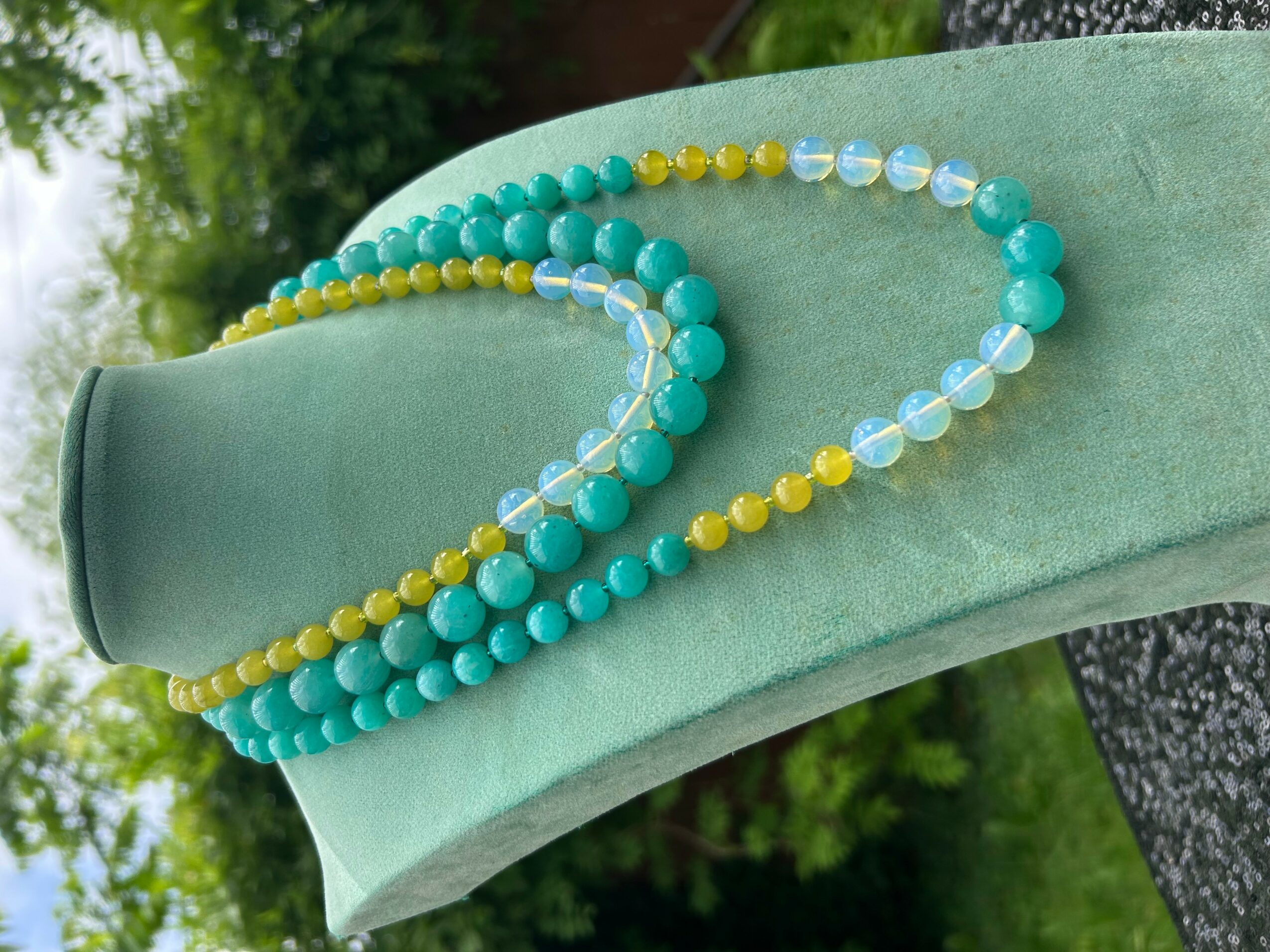 Two beaded necklaces, one with turquoise and yellow beads, draped over a green fabric surface outdoors.