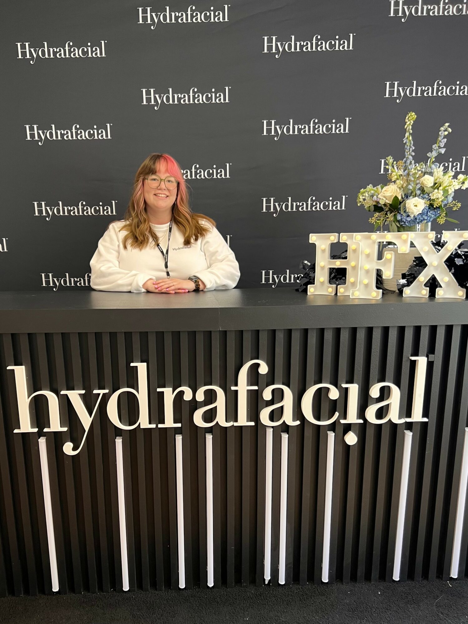 Woman with glasses and long hair sitting at a black reception desk with floral arrangement and large letters, backdrop with 'Hydrafacial' text.