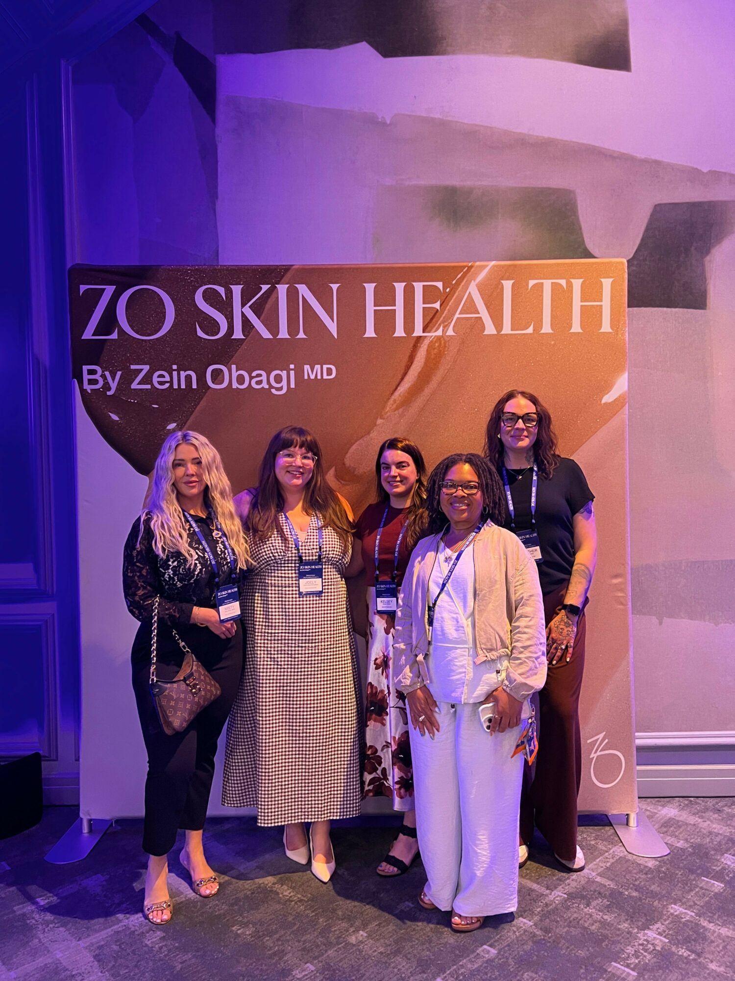 Group of six women standing in front of a health event banner, smiling, in an indoor setting.