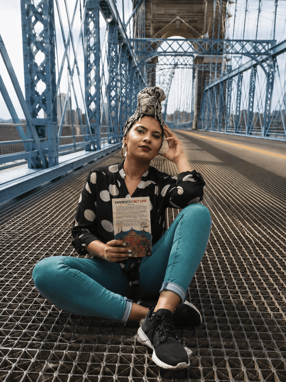 Woman sitting on a bridge with metal grating, holding a book, wearing a polka dot shirt and teal pants.