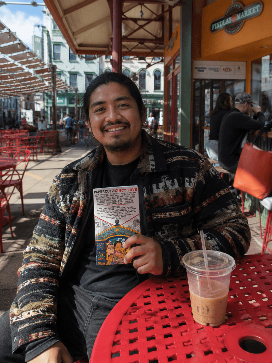 Man smiling at outdoor cafe, holding a menu, with a drink on red table, buildings and people in background.