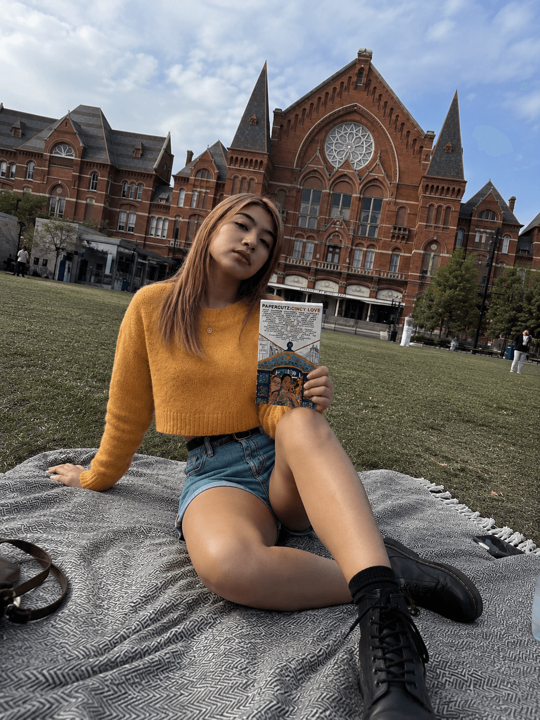 Young woman sitting on a blanket in front of a large historic brick building with pointed towers and a rose window, holding a phone.