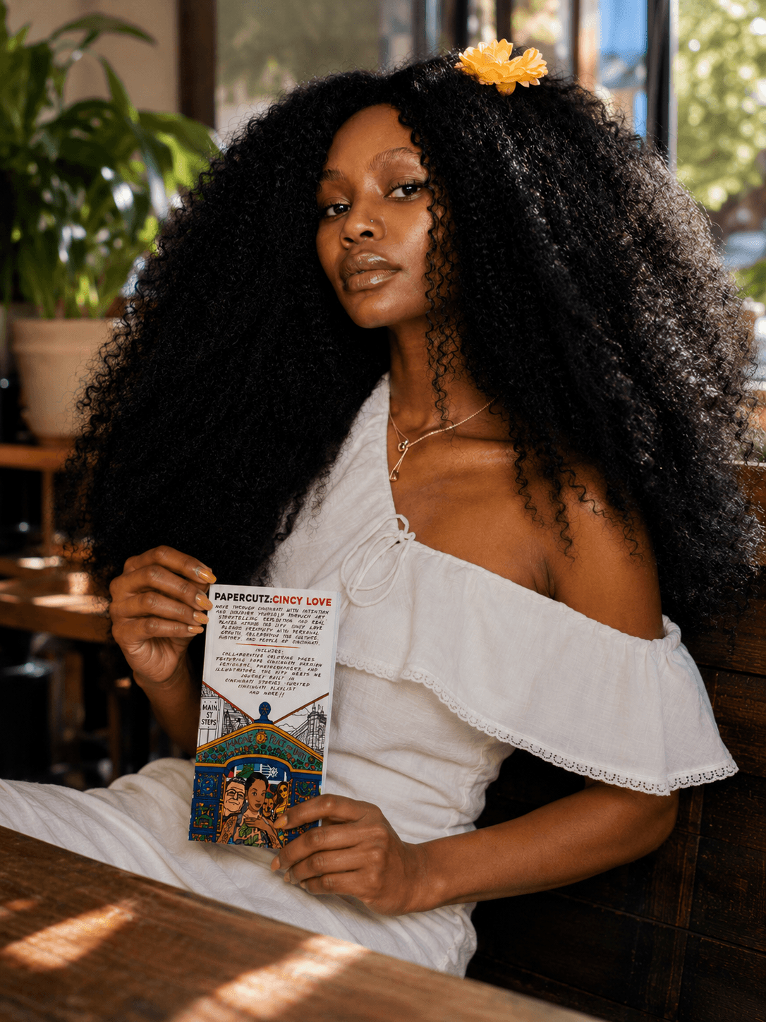 Woman with long curly hair wearing a white off-shoulder dress, holding a colorful card, sitting indoors with plants in background.