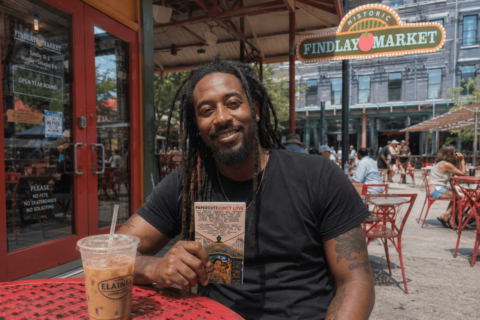 Man with dreadlocks smiling, holding a card, sitting outdoors at a market with red chairs and a sign reading 'Findlay Market'.