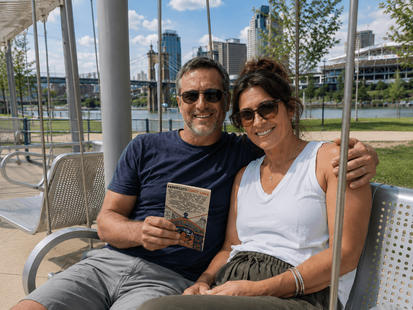 A smiling man and woman sit on a swing outdoors, with city buildings and a river in the background.