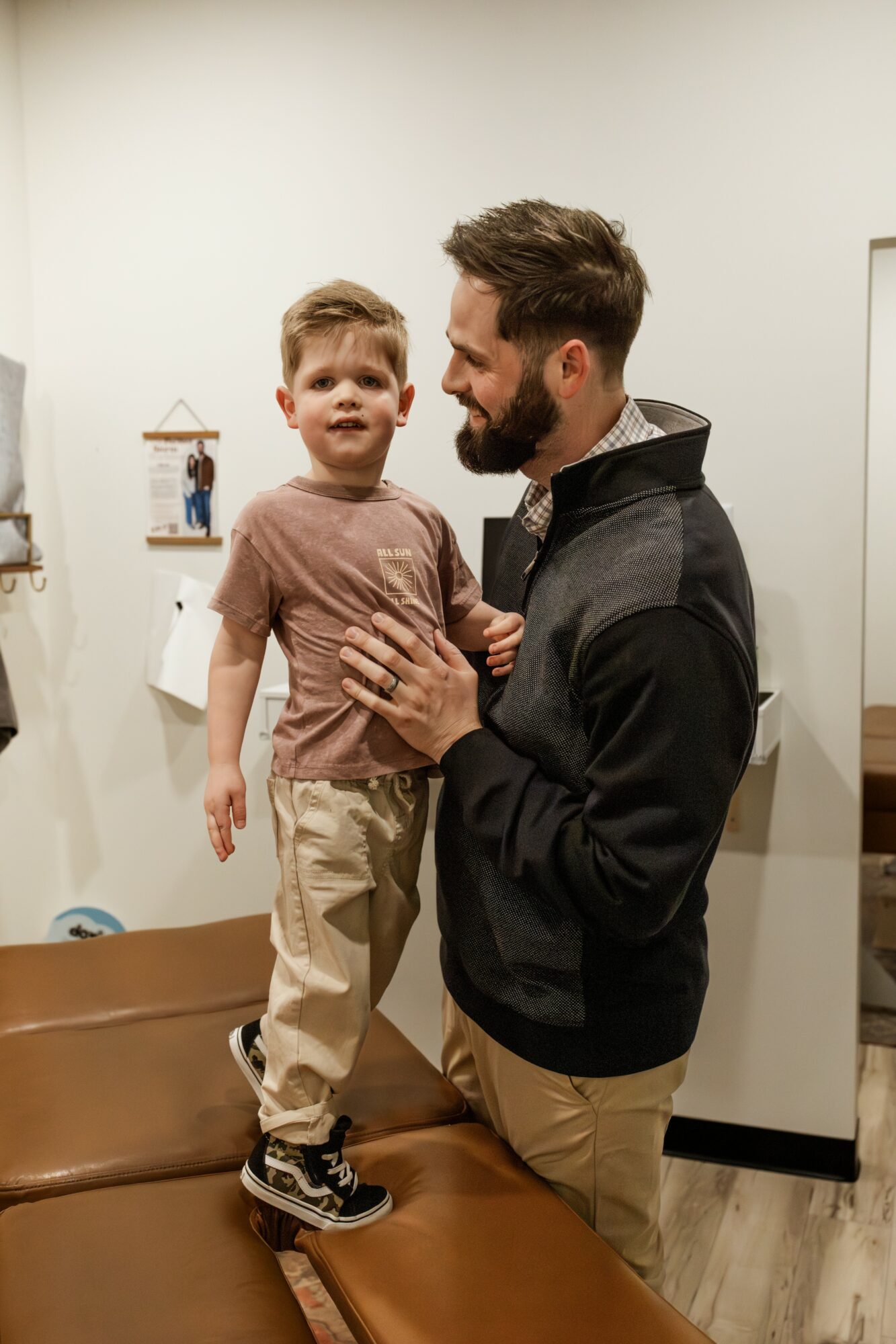 Man holding a young boy on a brown bench indoors, smiling and engaging with each other, plain wall background.