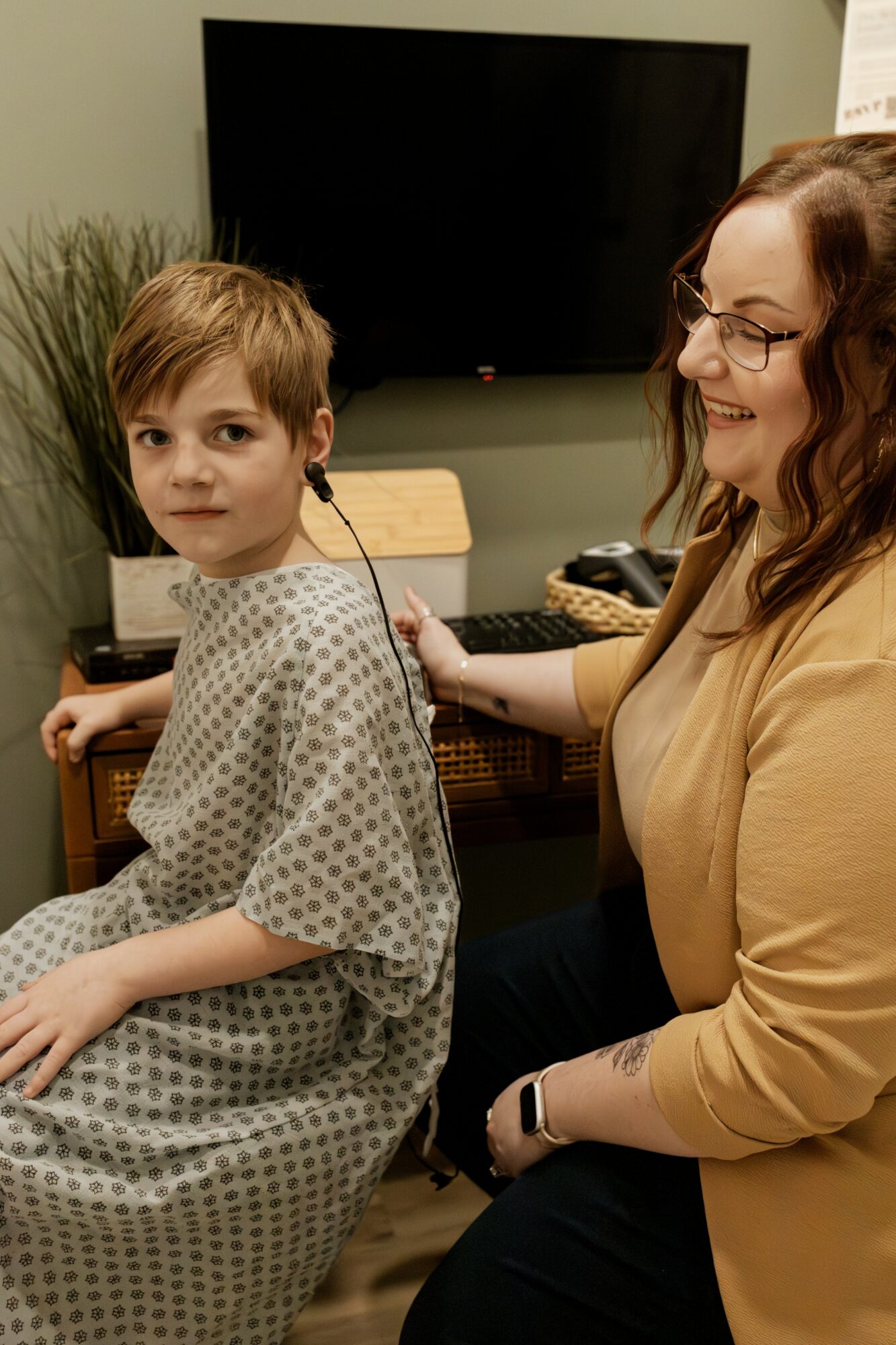 A woman with glasses and a young boy in a hospital gown sit together, smiling, in a room with a TV and plants.