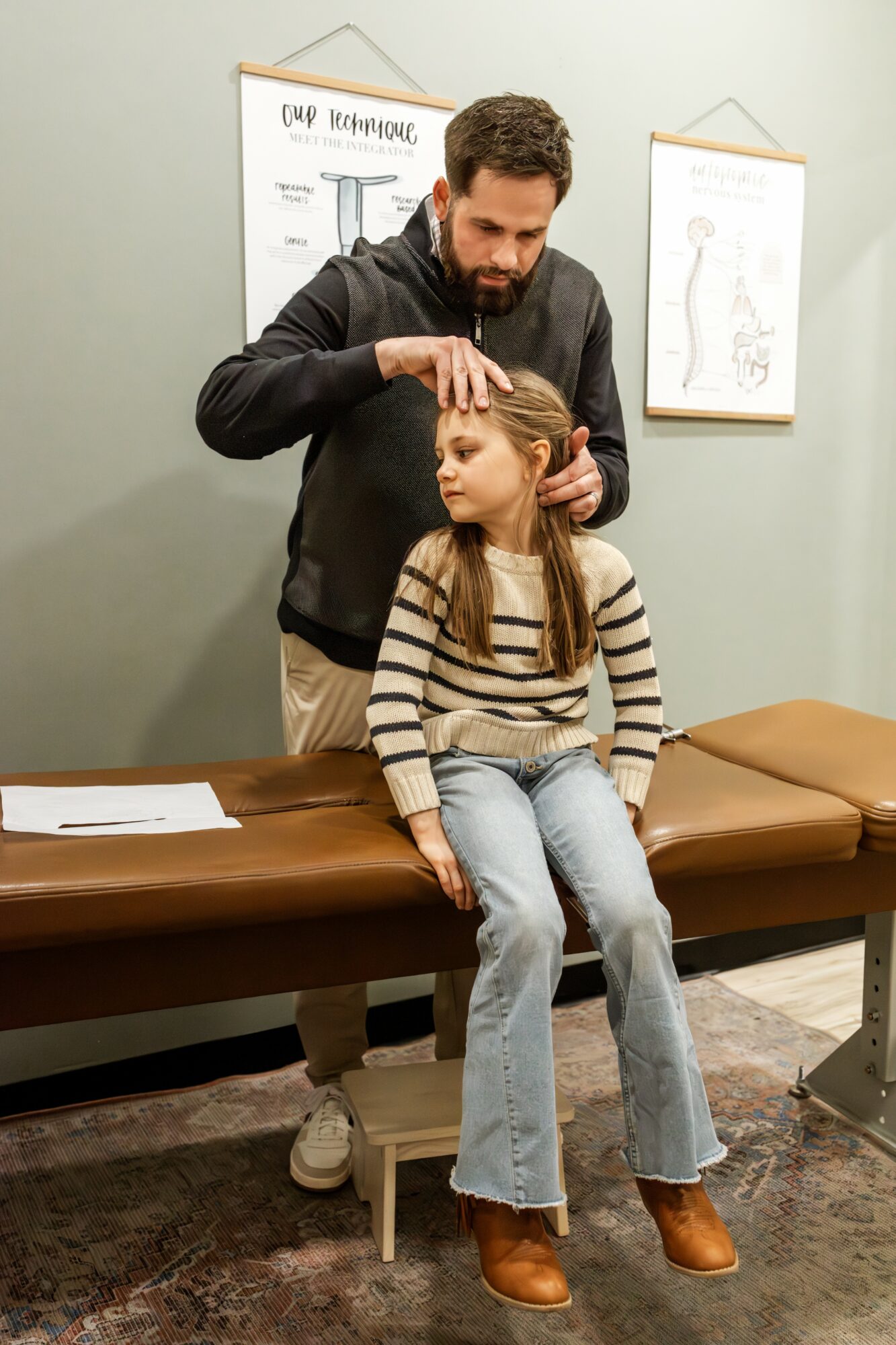 A man examines a young girl sitting on an examination table in a medical office, with medical posters on the wall behind them.