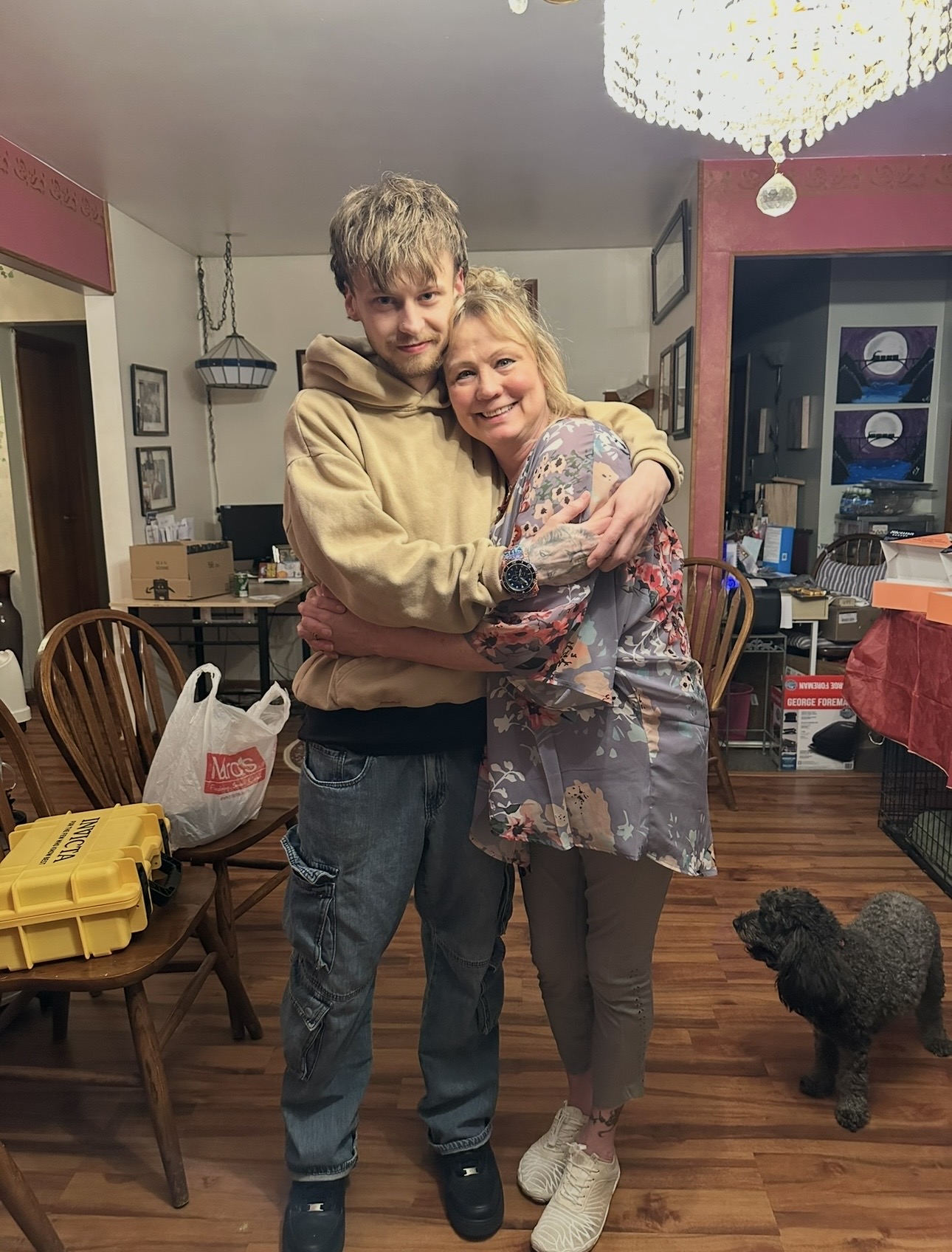 Two people hugging in a home, with a dog nearby and dining area in the background.