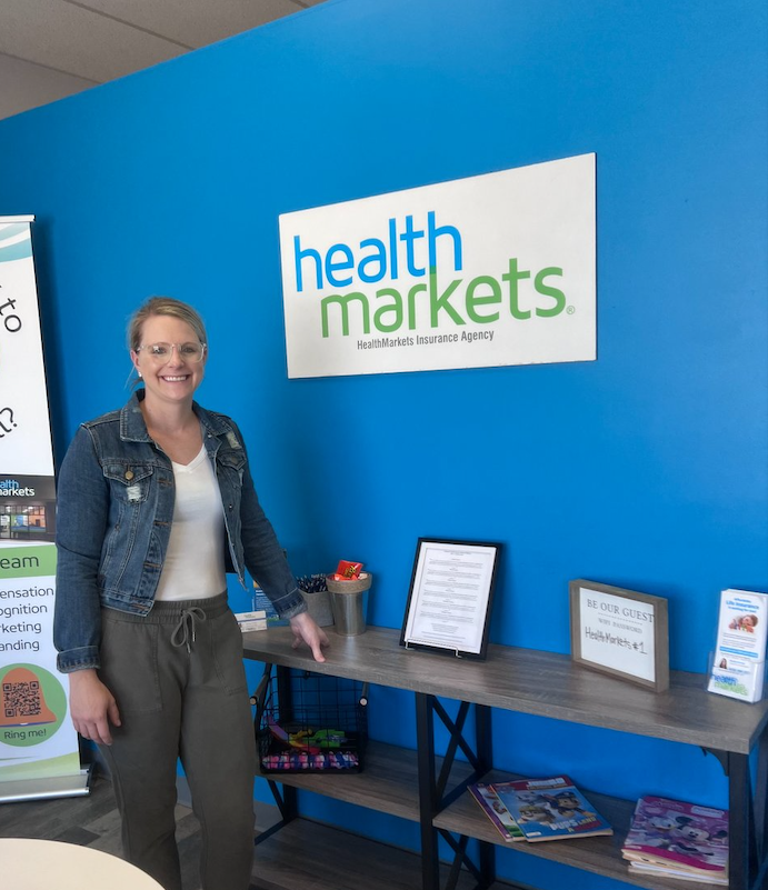 Smiling woman standing next to a table with magazines and framed signs in front of a blue wall with a 'health markets' sign.