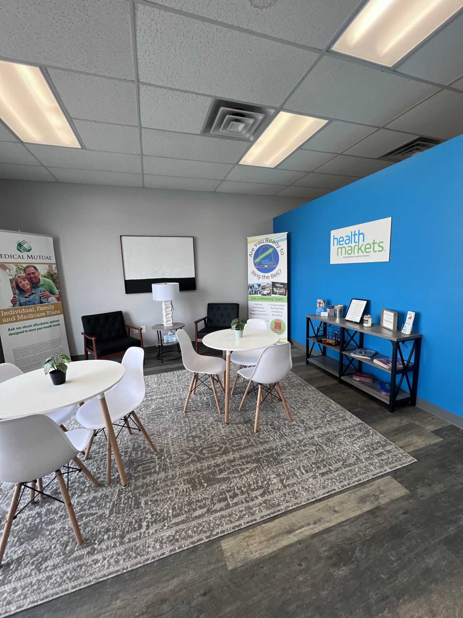 Indoor waiting area with round tables, chairs, a rug, and a blue wall with a sign reading 'health markets'.