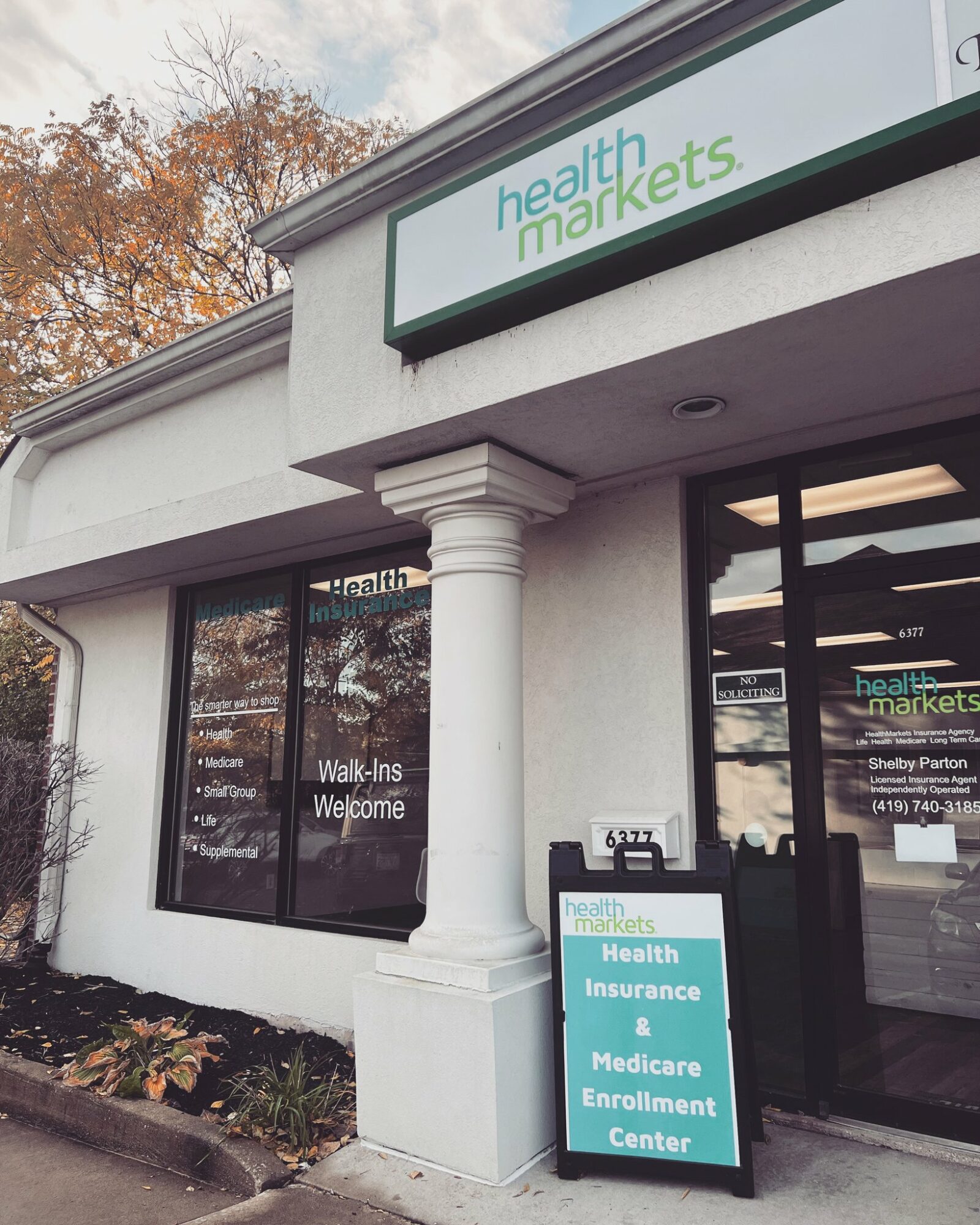 Exterior of a health insurance office with large windows and a sign for health markets, with a sidewalk and trees outside.