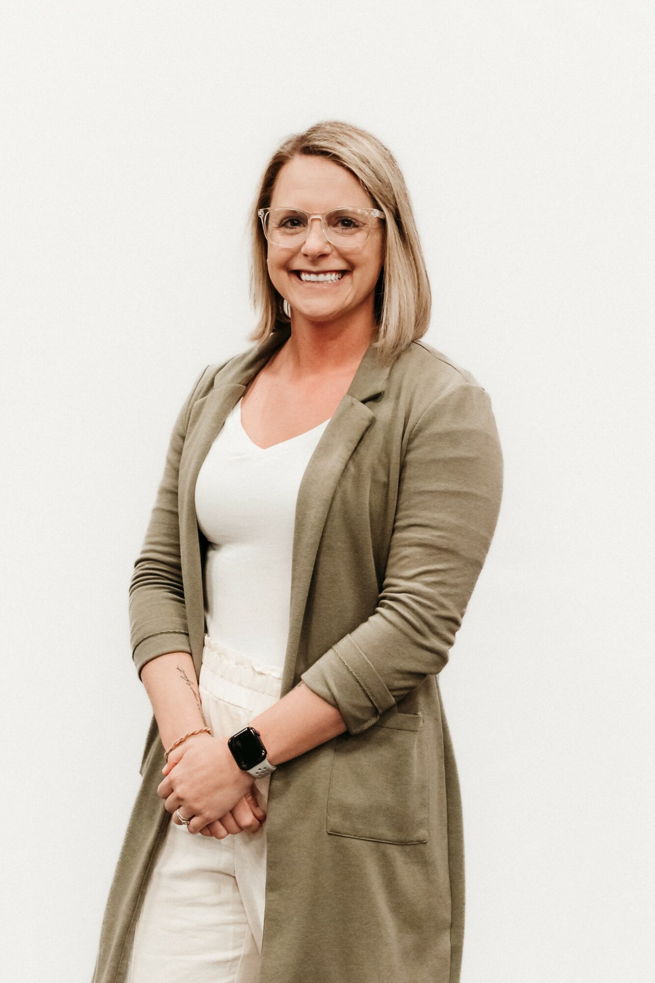 Woman with shoulder-length blonde hair, glasses, smiling, wearing a beige blazer over a white top, standing against a plain white background.